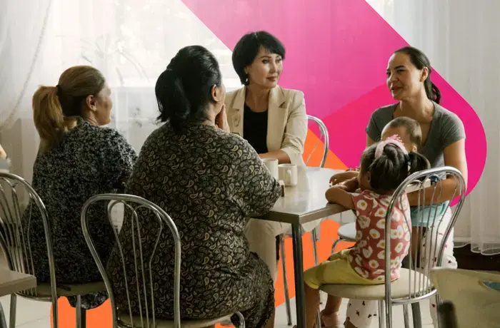 Four women sit and talk around a table. One woman holds a small child in her arms.