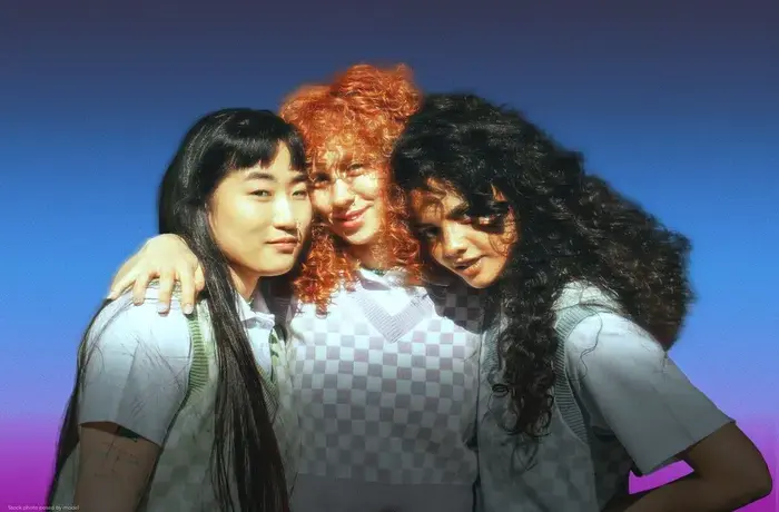Three young, diverse women standing close together with their arms around each other against a blue background with a pink gradient at the bottom. They are facing the camera with relaxed expressions and are wearing light-coloured tops layered with patterned sweater vests.