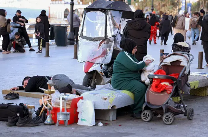 A woman sits beside a stroller caring for a child on a busy street, with a person lying nearby.