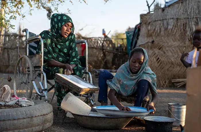A woman in a wheelchair sits outdoors beside a girl washing dishes, with a child standing nearby.