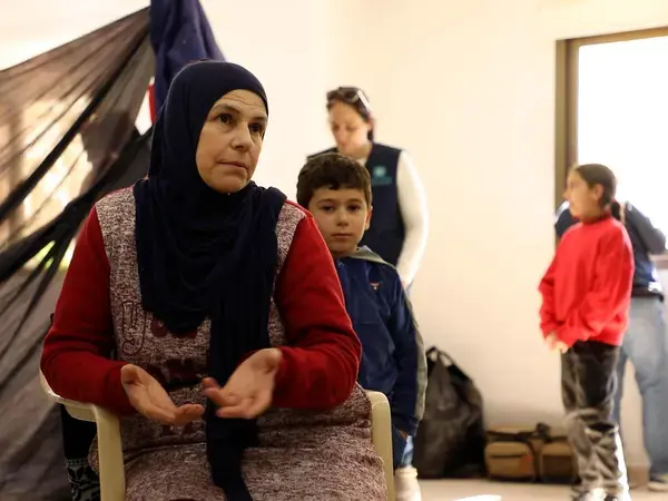 A woman gestures with her hands while seated indoors, with children and other people standing behind her.