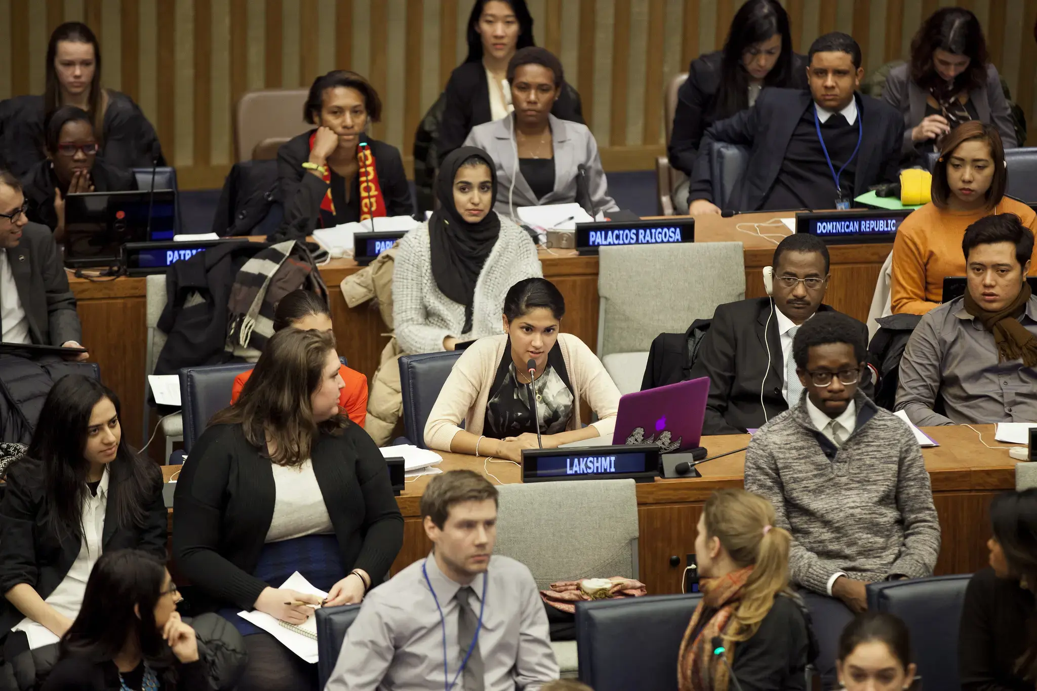 Youth participating in a youth forum inside a large conference hall. A young woman is speaking into a microphone from her seat, with a laptop open in front of her and a nameplate displayed. Other young people sit around her listening attentively, some taking notes or looking toward the speaker. Country nameplates are visible on desks, indicating a formal youth forum setting.