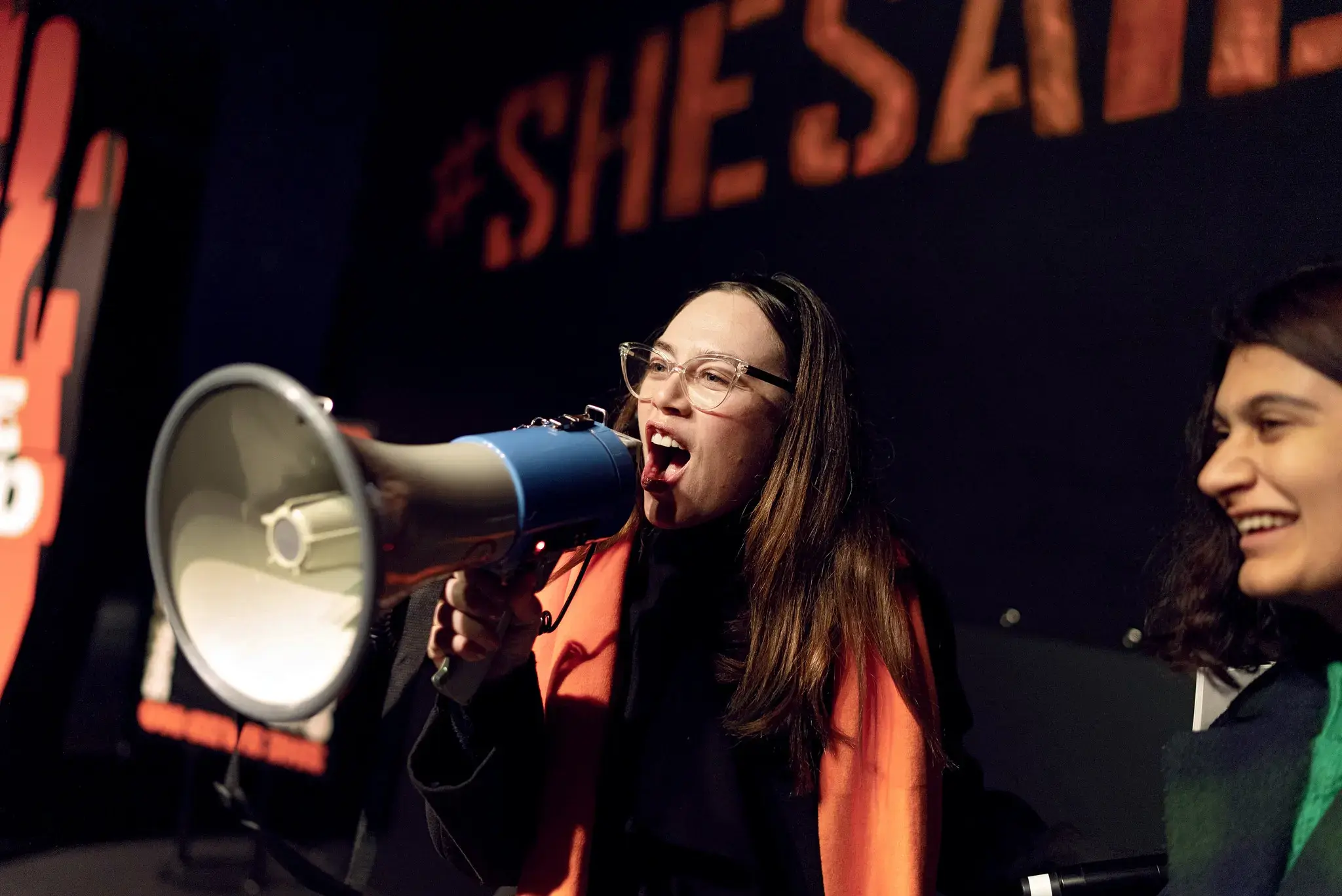 A woman speaks into a megaphone at a UN Women event, standing on a stage with a dark backdrop displaying the hashtag “#SheSaid” in orange. She is wearing glasses and an orange scarf over dark clothing. Another woman stands beside her smiling.