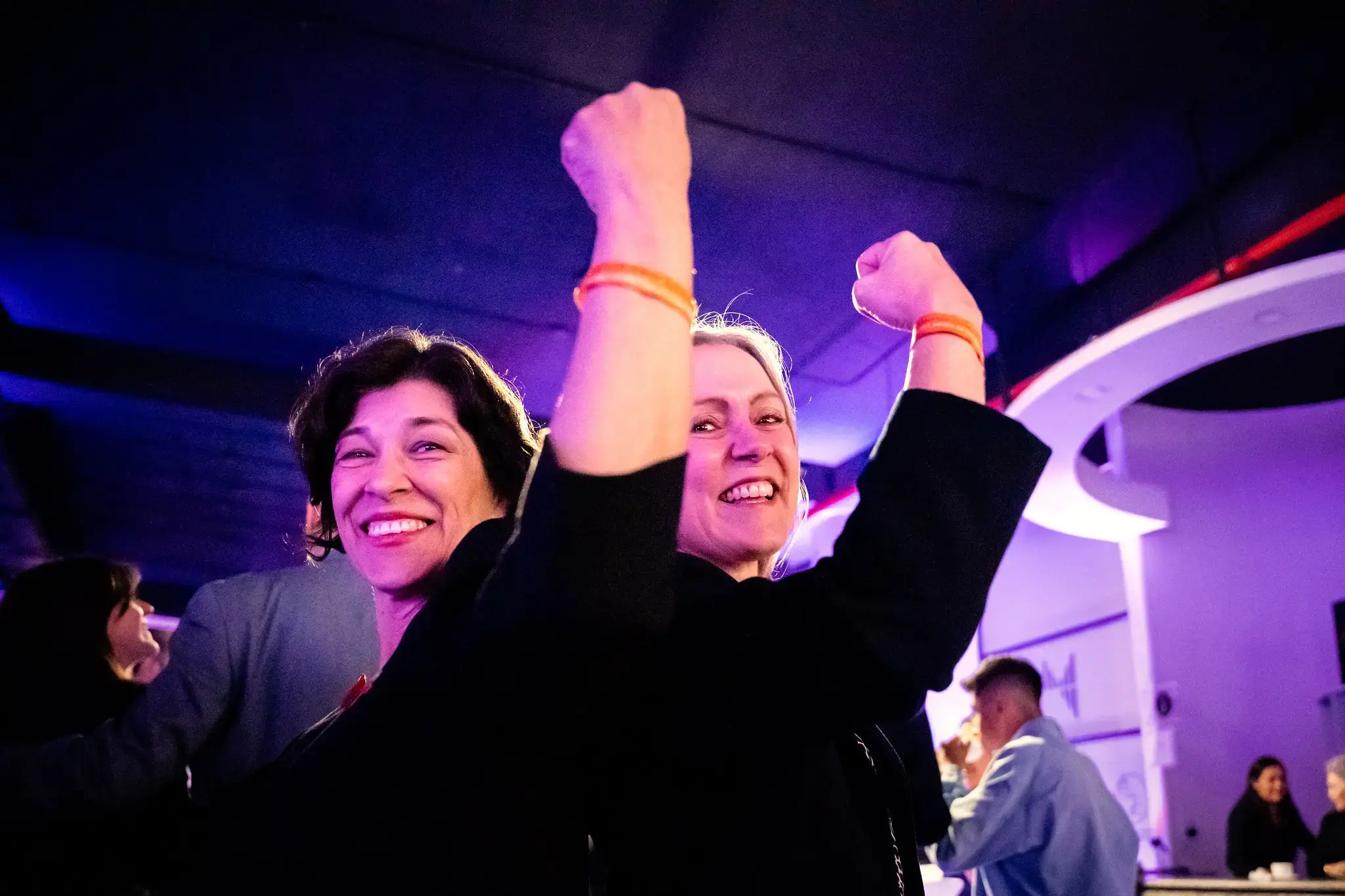 Two women smiling and raising their fists in celebration at a UN Women event. They stand close together under purple and blue event lighting, wearing dark clothing and orange wristbands. Other participants are visible in the background.