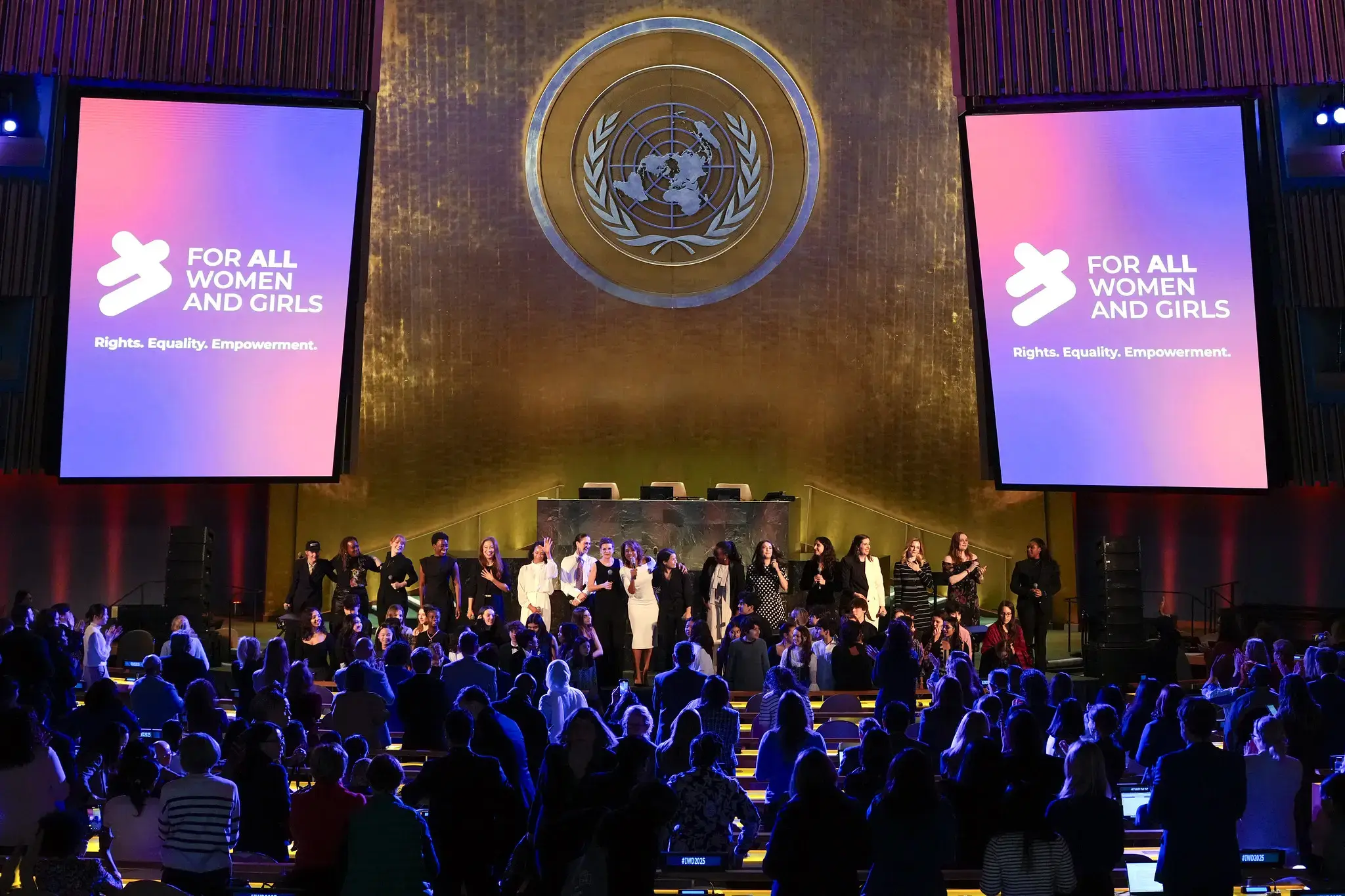 Wide view of the United Nations General Assembly Hall during an International Women’s Day event. Two large screens display the message “For All Women and Girls – Rights. Equality. Empowerment.” A group of women stand together on the stage beneath the UN emblem, while a large audience stands and applauds in the hall. The scene is lit with purple and blue lighting.