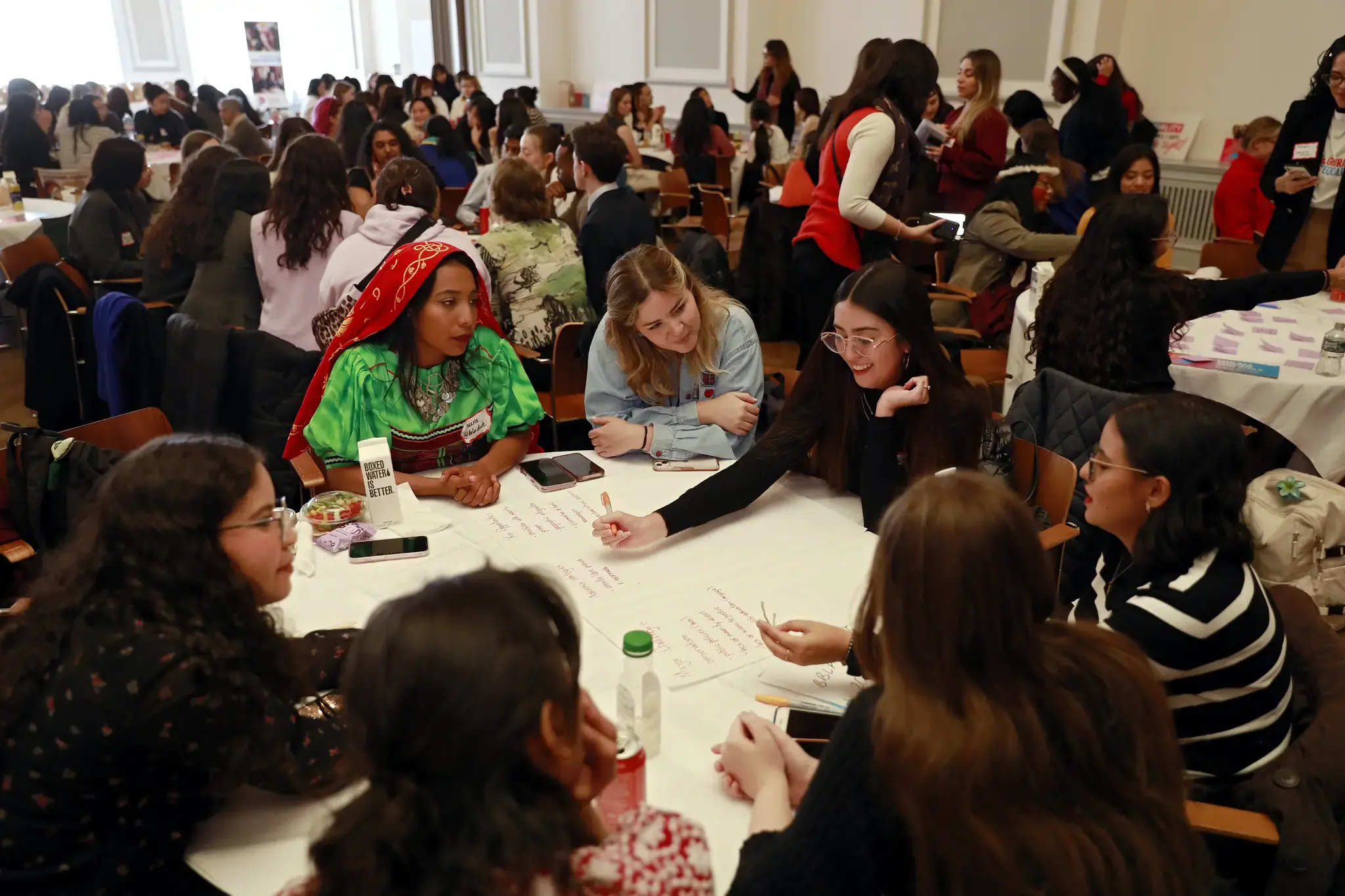 Young people participating in a youth dialogue at round tables in a conference room. A group of young women sit together writing notes on a large sheet of paper, discussing ideas and smiling. Other youth groups are visible in the background engaged in similar discussions, creating an active and collaborative atmosphere.