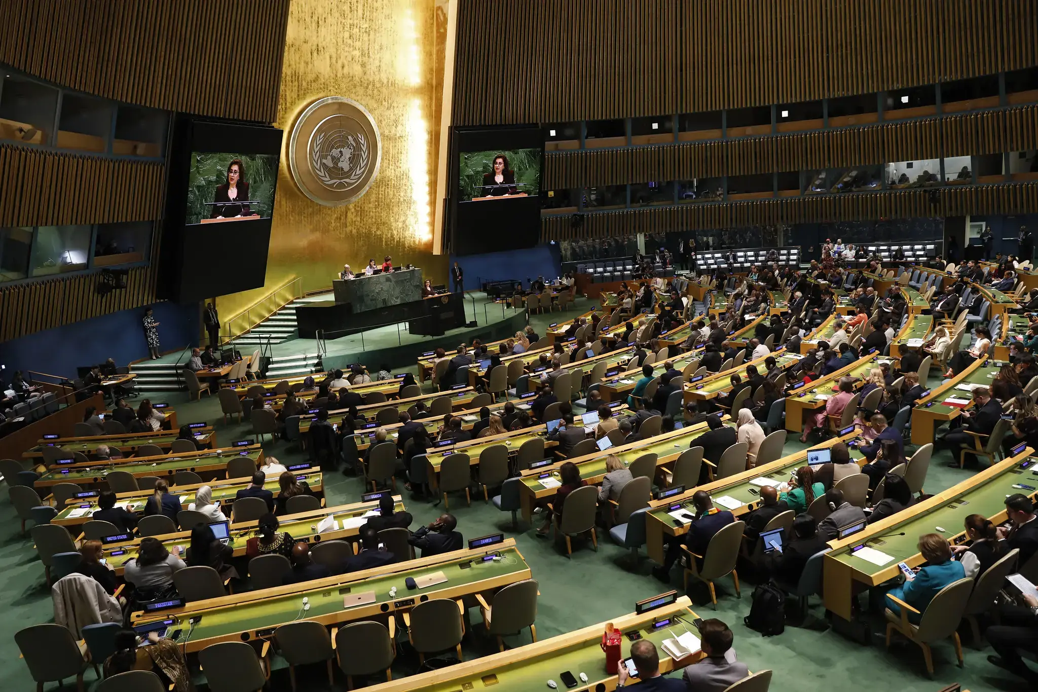 Wide view of the United Nations General Assembly Hall during a formal meeting. UN Women Executive Director Sima Bahous is addressing the assembly from the main podium, with her image displayed on two large screens. Delegates are seated in curved rows facing the dais, with the UN emblem visible on the wall behind her. Participants sit at desks with microphones, nameplates and laptops, listening and taking notes.